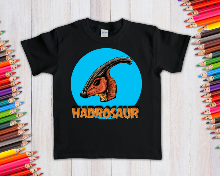 The Kids Hadrosaur Head T-Shirt by Jurassic Studio features a vibrant parasaurolophus head in a blue circle above HADROSAUR, surrounded by colored pencils—a great dinosaur fan gift, shown on a light wooden background.