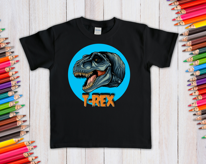 Jurassic Studio’s Kids T-Rex Head T-Shirt features a cartoon dino head in a blue circle with T-REX below on black, shown on wood with colorful pencils—an exclusive gift perfect for young dinosaur fans.