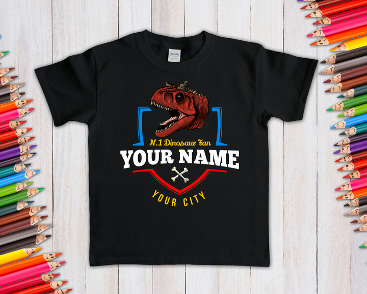 Jurassic Studio’s N.1 Carnotaurus Fan Custom Kids T-Shirt—black, features a red dinosaur and personalized “No. 1 Dinosaur fan YOUR NAME YOUR CITY” text; displayed with colorful pencils—perfect for dino-loving kids!.
