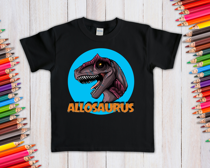 The Kids Allosaurus Head T-Shirt by Jurassic Studio features a bold dino head in a blue circle with orange text—displayed on a white wooden surface with colored pencils—making it the perfect gift for young dinosaur fans.