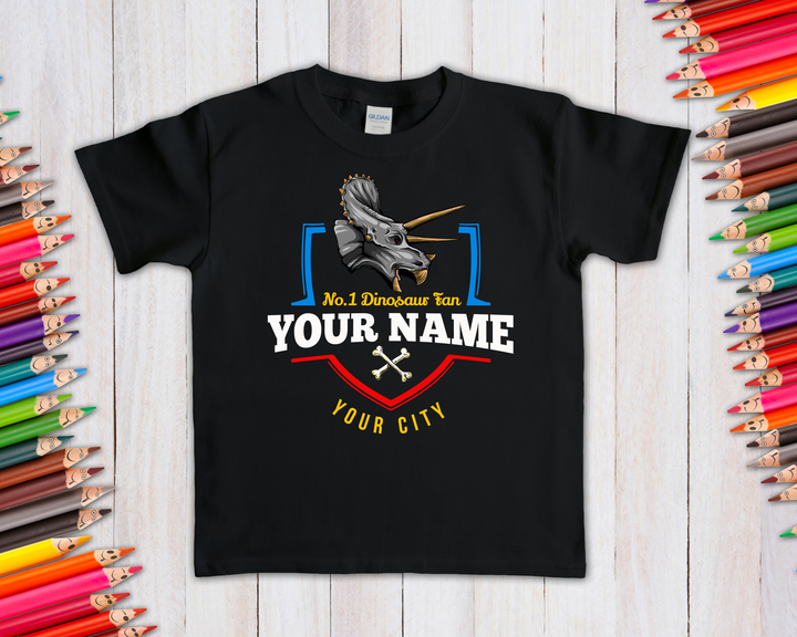Jurassic Studio’s N.1 Triceratops Fan Custom Kids T-Shirt features a triceratops head, No. 1 Dinosaur Fan text, your child’s name & city, small crossed bones, and displays on a wooden background with colored pencils.