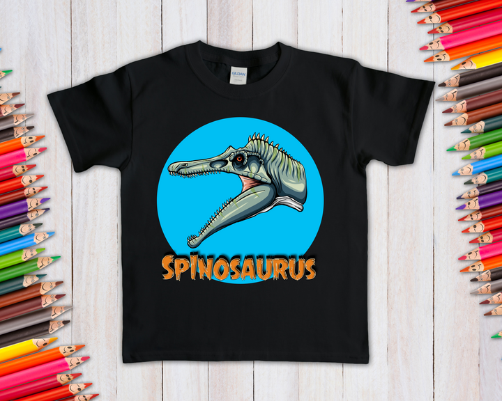 The Jurassic Studio Kids Spinosaurus Head T-Shirt features a cartoon dino head in a blue circle with SPINOSAURUS in orange below, shown on a light wooden background with colored pencils around it.