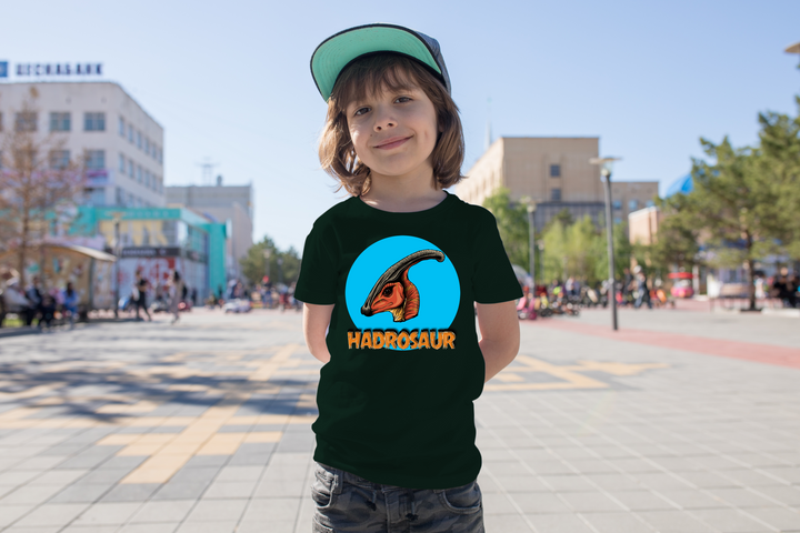 A smiling child wears the black Kids Hadrosaur Head T-Shirt by Jurassic Studio, featuring a colorful dinosaur graphic—an ideal gift for dinosaur fans. City buildings and people fill the background.