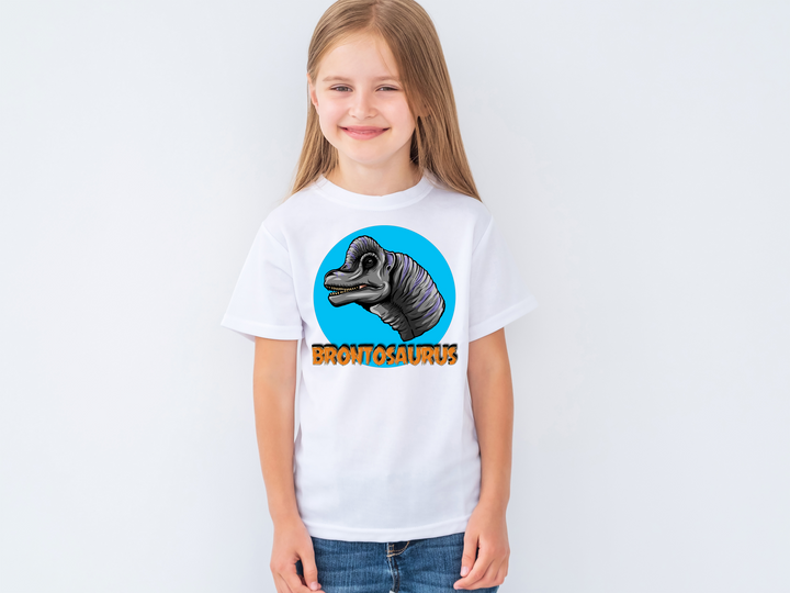 A young girl with long blonde hair wears the Jurassic Studio Kids Brontosaurus Head T-Shirt and smiles against a plain light background—an ideal dinosaur fan gift for any young enthusiast.