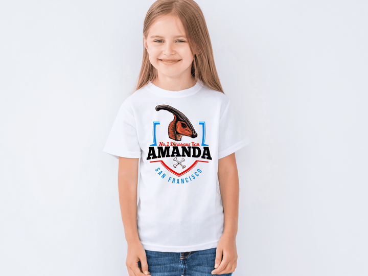 Smiling young girl with long light brown hair wears the Jurassic Studio N.1 Hadrosaur Fan Custom Kids T-shirt—a white tee featuring a Parasaurolophus, No. 1 Dinosaur Fan Amanda, San Francisco, and two small crossed bones.