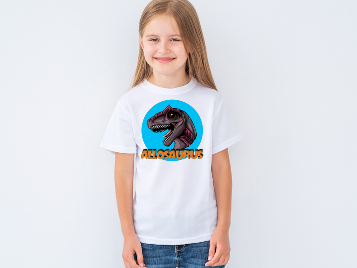 A young girl smiles in a Jurassic Studio Kids Allosaurus Head T-Shirt, showing a vibrant Allosaurus head illustration and the word ALLOSAURUS—a perfect gift for any dinosaur fan.