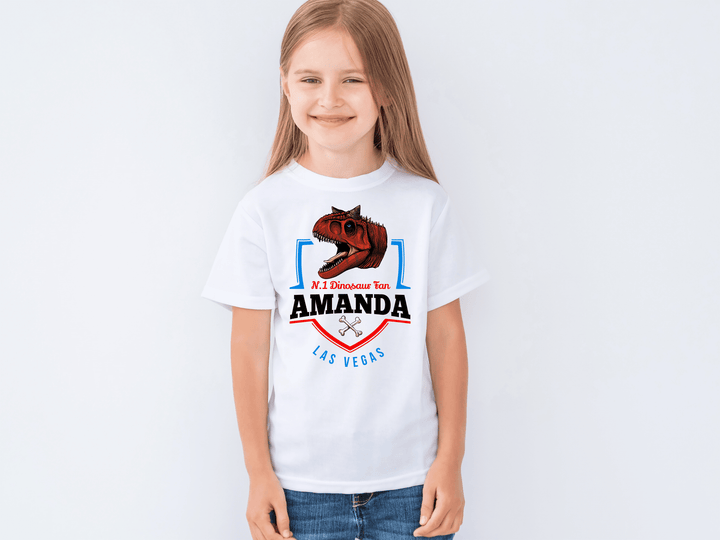 A smiling young girl with long light brown hair wears the N.1 Carnotaurus Fan Custom Kids T-Shirt by Jurassic Studio, featuring a roaring dinosaur, her name Amanda, and “Mi Dinosaur Tour Las Vegas,” standing against a plain white background.