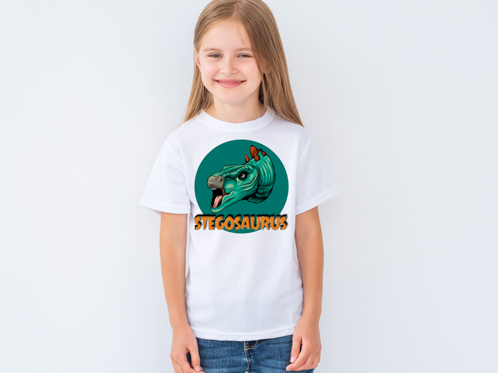 A smiling young girl models the Jurassic Studio Kids Stegosaurus Head T-Shirt, featuring a cartoon stegosaurus head and STEGOSAURUS below—an ideal gift for dinosaur fans, shown against a plain light background.