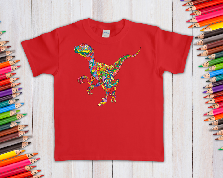 The Jurassic Studio Kids Velociraptor Zentangle T-Shirt features a vibrant dinosaur design, shown flat on a white wooden surface with colored pencils—perfect gift for any young dino fan.