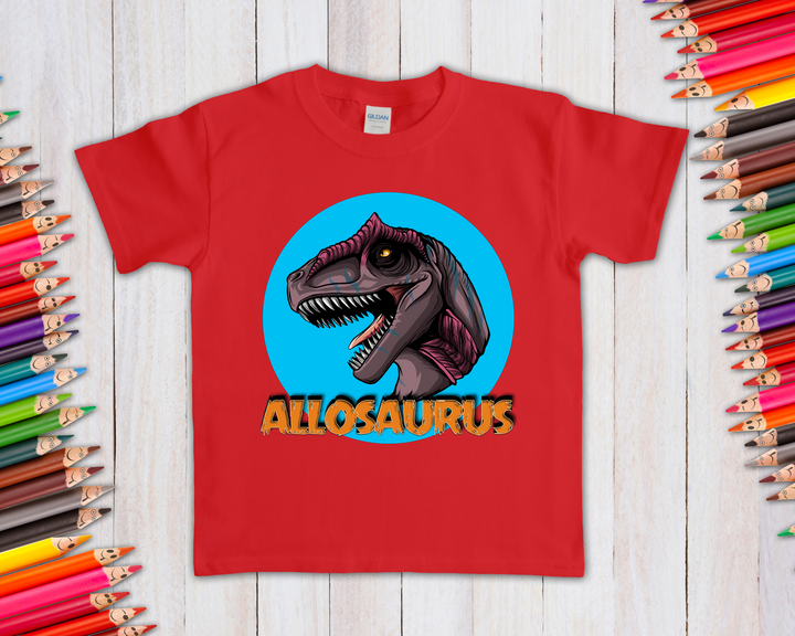 The Jurassic Studio Kids Allosaurus Head T-Shirt features a red shirt with an illustrated Allosaurus head in a blue circle above bold orange text, shown on a light wooden background with colorful pencils—perfect for any dinosaur fan.