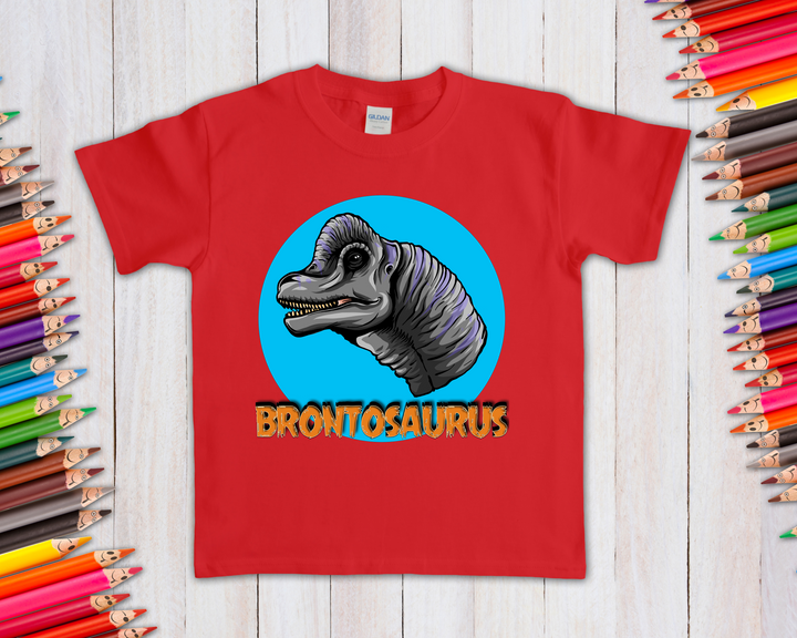 The Jurassic Studio Kids Brontosaurus Head T-Shirt showcases a cartoon brontosaurus in a blue circle with bold yellow text and colorful pencils—an ideal gift for dinosaur fans. Displayed on a white wooden surface.