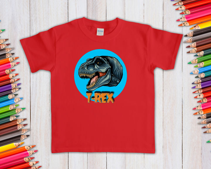 The Jurassic Studio Kids T-Rex Head T-Shirt showcases a roaring dinosaur in a blue circle with T-REX text—an ideal gift for dinosaur fans. Displayed on a white wooden surface, its framed by colorful pencils.