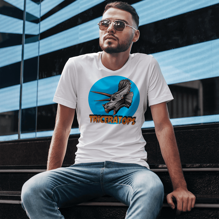 A man with short hair and sunglasses sits on steps in jeans, wearing the Jurassic Studio Men Triceratops Head T-Shirt featuring a triceratops in a blue circle with “TRICERATOPS” below—an ideal gift for dinosaur fans.