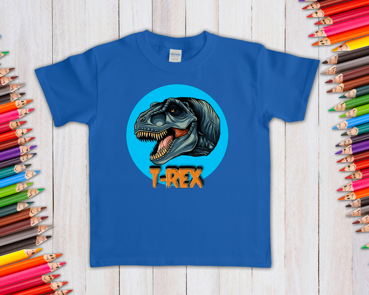 The Jurassic Studio Kids T-Rex Head T-Shirt features a snarling dino head in a blue circle with bold orange T-REX text below. Perfect for dinosaur fans and shown with colored pencils on a light wooden surface.