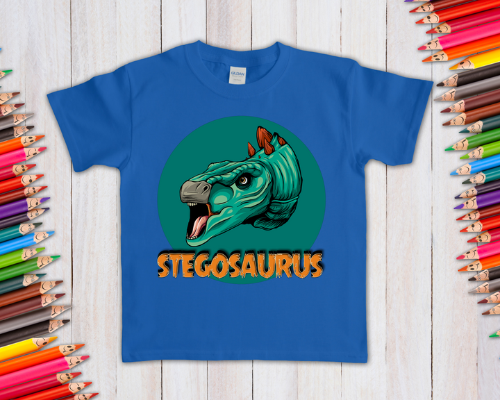 The Kids Stegosaurus Head T-Shirt by Jurassic Studio features a green and orange illustrated stegosaurus head above bold STEGOSAURUS text—a perfect dinosaur fan gift. Colored pencils border the wooden background.