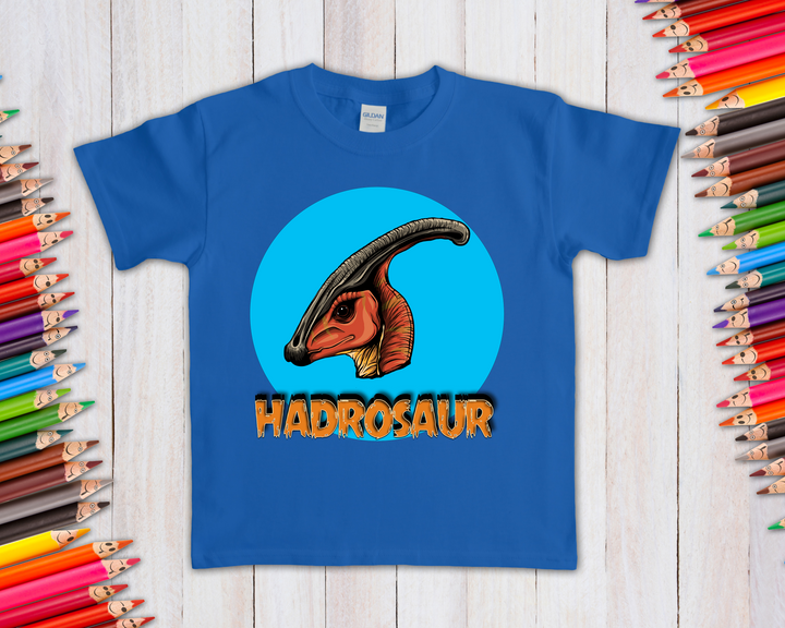 The Kids Hadrosaur Head T-Shirt by Jurassic Studio showcases an illustrated hadrosaur head in a blue circle with bold orange HADROSAUR text—ideal for dinosaur fans. Displayed with colored pencils on a light wooden surface.