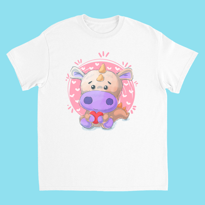 Jurassic Studio’s Kids Pink Baby Dino T-Shirt: a white cotton tee with a cute cartoon dino holding a red heart, set in a pink circle with small hearts on a light blue background.