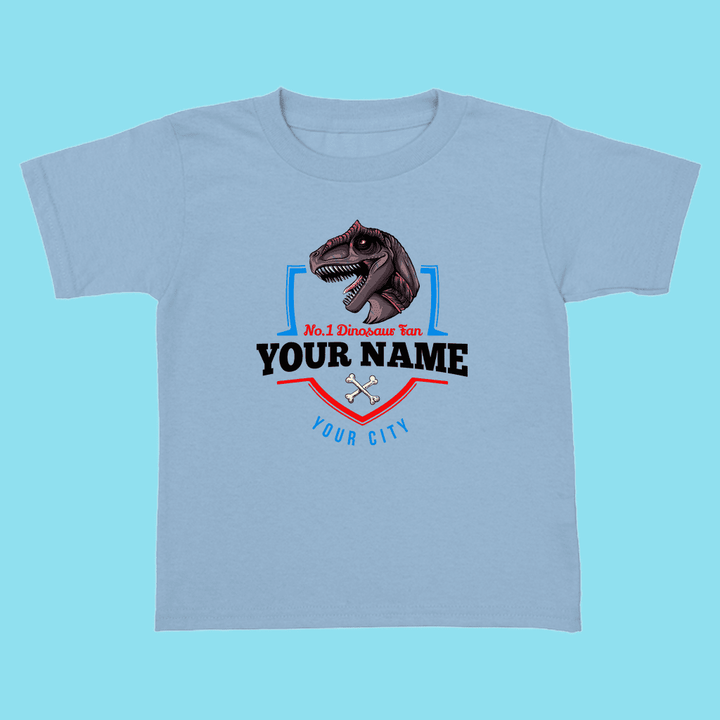 The Jurassic Studio N.1 Allosaurus Fan Custom Toddler T-Shirt is a light blue kids’ shirt with a bold Allosaurus head, red and blue shield accents, crossed bones, and custom “No.1 Dinosaur Fan, YOUR NAME, YOUR CITY” text.