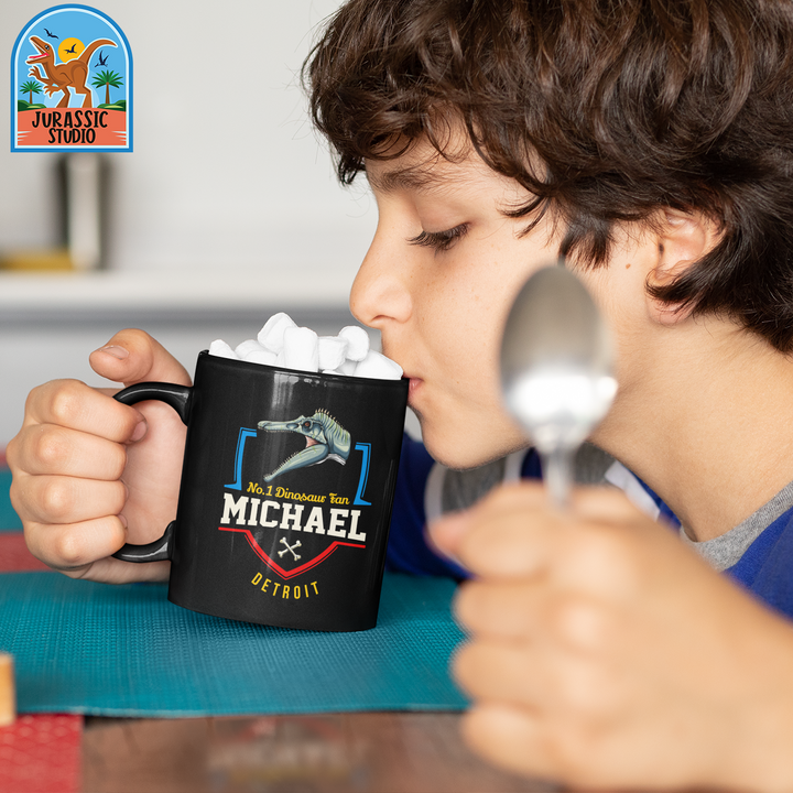 A boy holds a spoon and drinks from a black N.1 Spinosaurus Fan Custom Mug personalized with Michael, Detroit, and a dinosaur graphic. This Jurassic Studio mug is topped with marshmallows, making a perfect dinosaur gift.