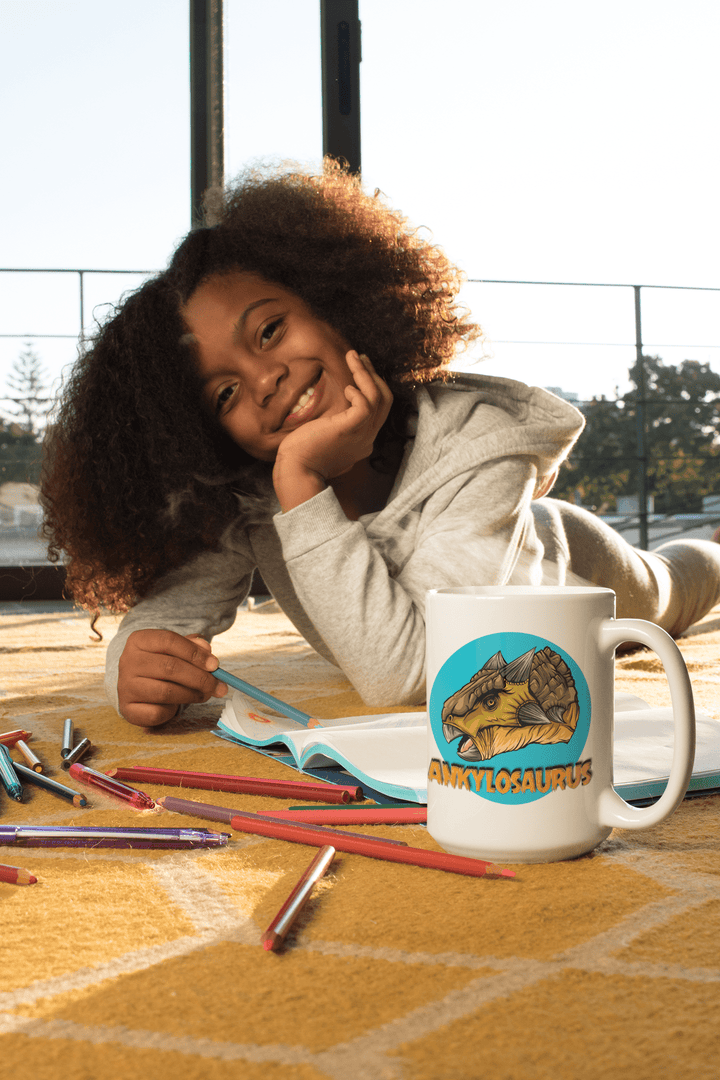 A smiling child lies on a yellow rug with colored pencils and an open notebook, next to the Jurassic Studio Ankylosaurus Portrait Mug, a white ceramic mug featuring a cartoon Ankylosaurus. Sunlight streams in from the window behind her.