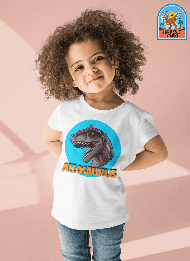 A young child with curly hair smiles, wearing the Jurassic Studio Toddler Allosaurus Head T-Shirt featuring a purple dinosaur and bold ALLOSAURUS text—perfect as a dinosaur fan gift. Light pink background includes the Jurassic Studio logo.