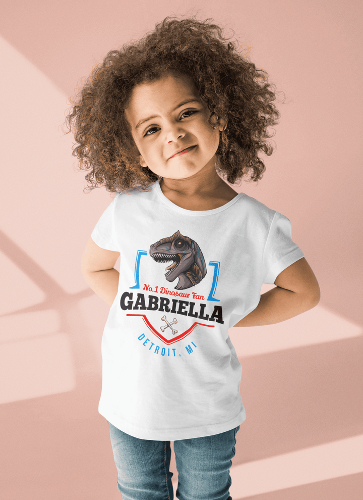 A curly-haired girl smiles with hands on hips, wearing the Jurassic Studio N.1 Allosaurus Fan Custom Toddler T-Shirt personalized with No. 1 Dinosaur Fan Gabriella Detroit, MI on a light pink background.