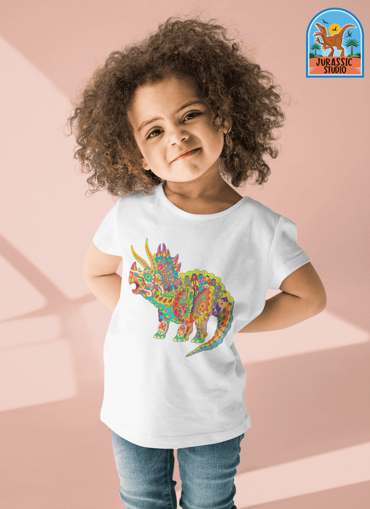 A smiling child with curly hair wears the Jurassic Studio Toddler Triceratops Zentangle T-Shirt featuring a vibrant dinosaur design. Set against a soft pink background, the Jurassic Studio logo appears in the upper right—a great gift for dino fans.