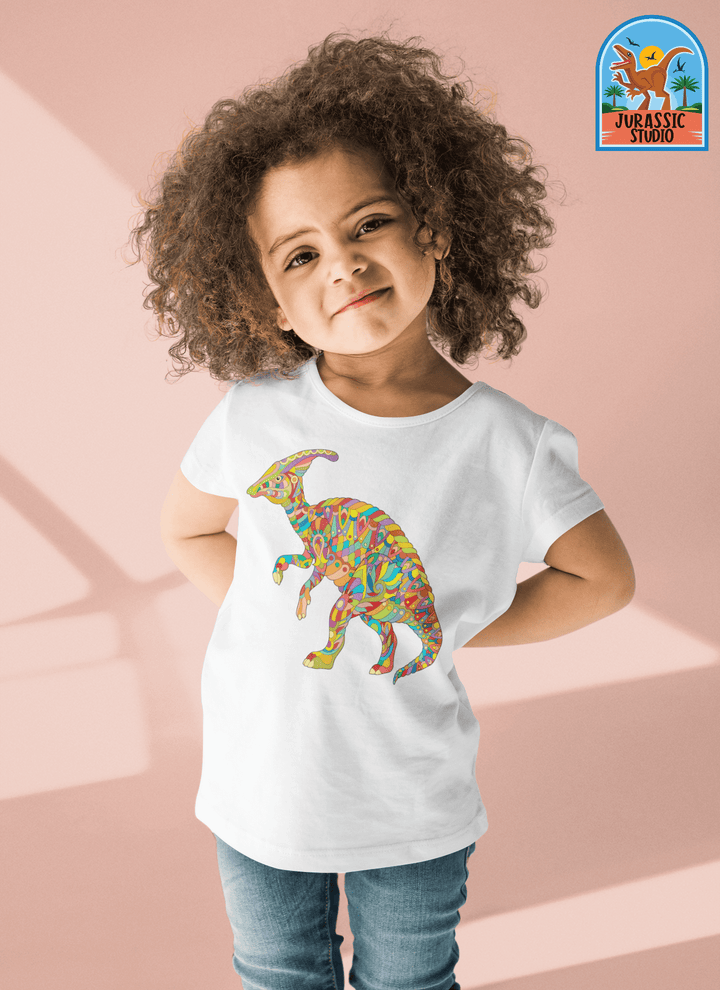 A young child with curly hair smiles, hands on hips, wearing the Toddler Hadrosaur Zentangle T-Shirt by Jurassic Studio—a 100% cotton tee with a vibrant dinosaur graphic. The Jurassic Studio logo is in the top right corner. Perfect for little dino fans!.
