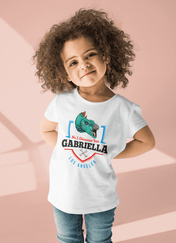 A young child with curly hair smiles, wearing Jurassic Studio’s N.1 Stegosaurus Fan Custom Toddler T-Shirt in pink—personalized with “No.1 Dinosaur Fan Gabriella, Los Angeles”—a perfect dinosaur gift for toddlers.