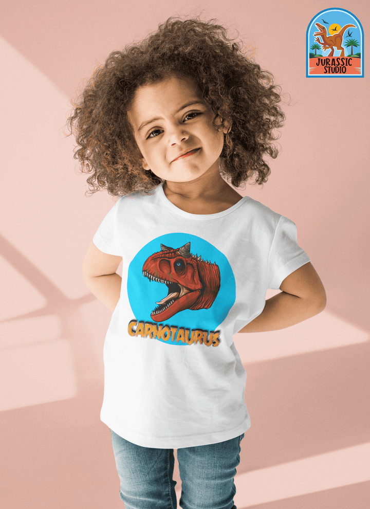 A smiling young child with curly hair poses in a Jurassic Studio Toddler Carnotaurus Head T-Shirt—a white tee featuring a red Carnotaurus dinosaur head in a blue circle, “CARNOTAURUS” below, and the Jurassic Studio logo at the top right.
