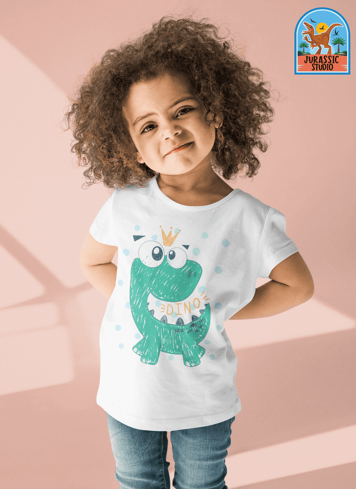 A young child with curly hair smiles confidently in a Jurassic Studio Toddler Baby Green Dino T-Shirt, featuring a cute green cartoon dinosaur and crown, set against a pale pink background. The Jurassic Studio logo appears in the top right corner.