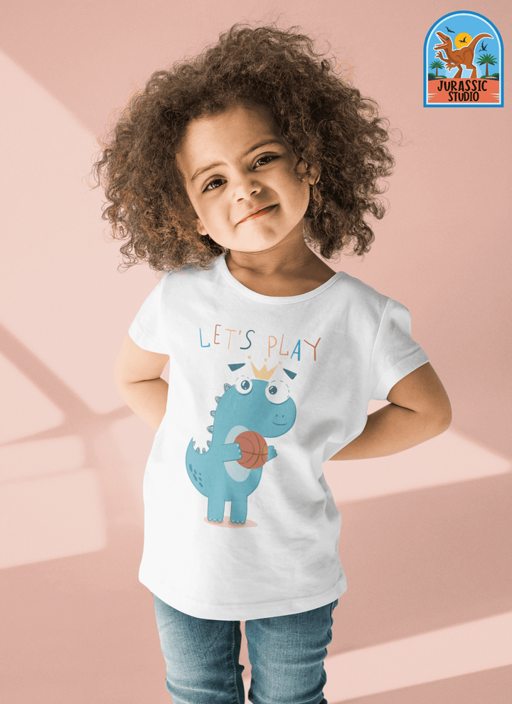 A smiling young child with curly hair wears the Toddler Baby Dino Basketball T-Shirt from Jurassic Studio, featuring a blue cartoon dinosaur with a basketball and LETS PLAY text. This exclusive design displays the Jurassic Studio logo in the upper right corner.