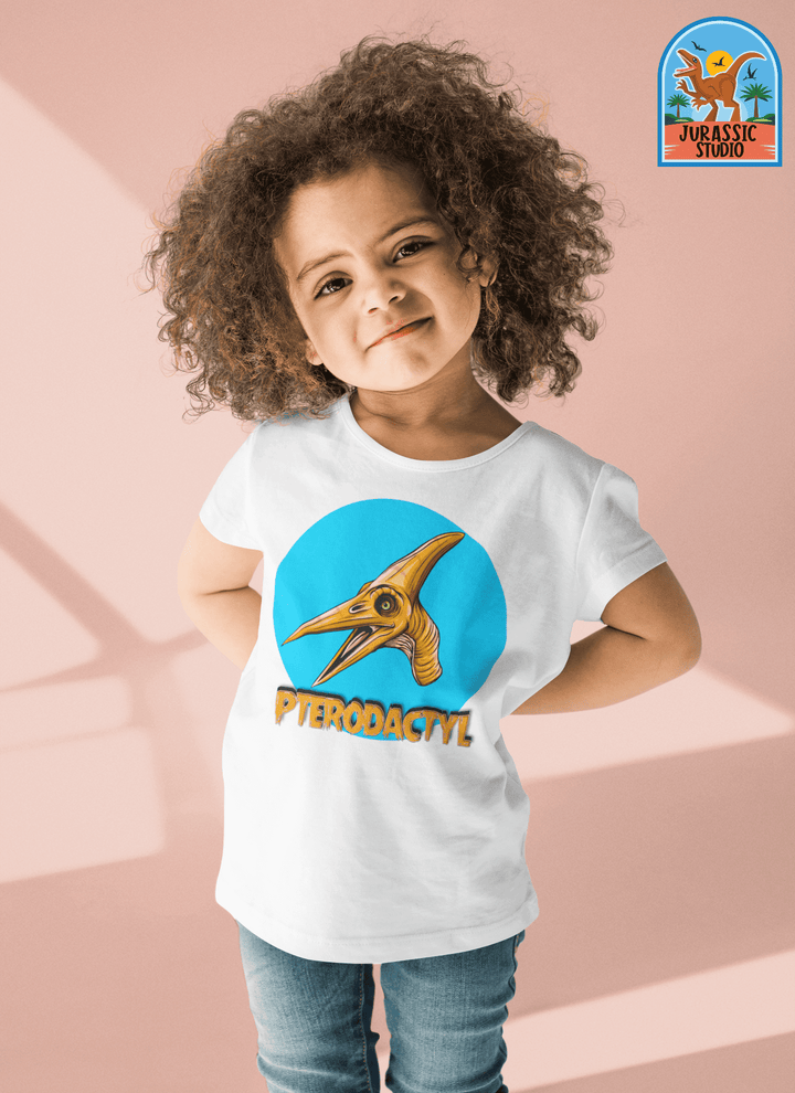 A smiling toddler with curly hair stands confidently in the Jurassic Studio Toddler Pterodactyl Head T-Shirt, featuring a cartoon pterodactyl on a bold blue circle with the brand logo at the top right—perfect for little dinosaur fans.