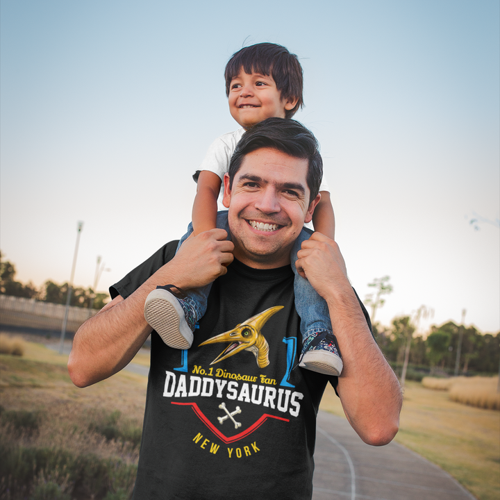 A smiling man carries a happy child on his shoulders outdoors at sunset, wearing a black Jurassic Studio N.1 Pterodactyl Fan Custom Men T-Shirt. The scene is set on a park path, capturing joyful family moments.