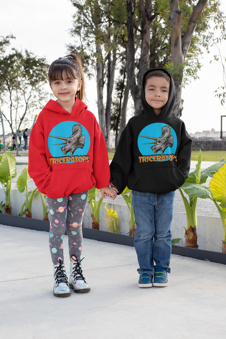 Two kids hold hands outdoors by plants, each in a Jurassic Studio Kids Triceratops Head Hoodie—one girl in red with leggings and one boy in black with jeans. Both hoodies feature playful triceratops graphics.