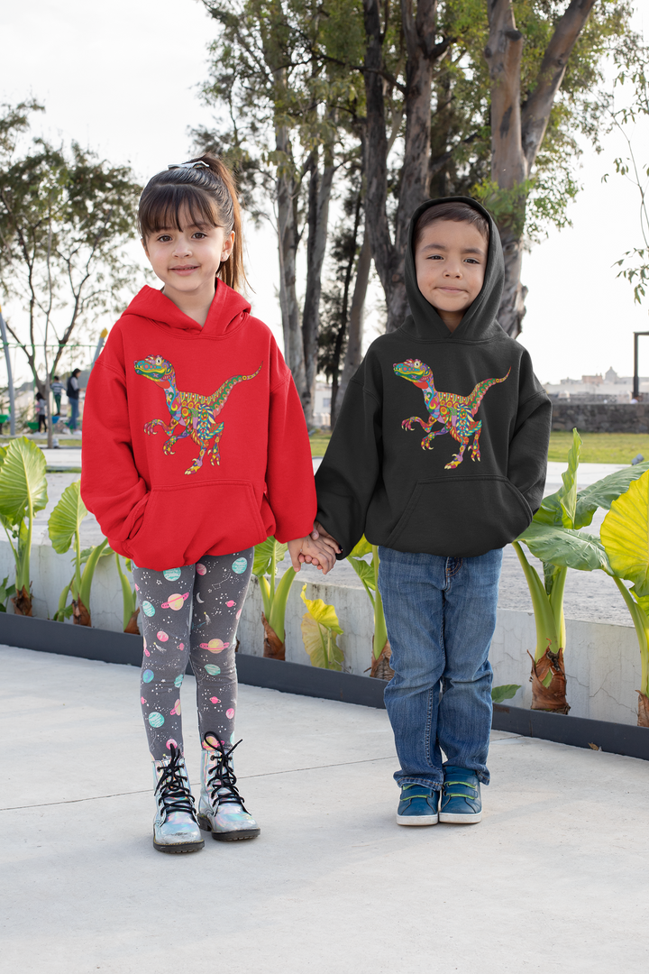 Two kids smile and hold hands outdoors by greenery, both in colorful Jurassic Studio Kids Velociraptor Zentangle Hoodies—the girl wears red with leggings, the boy wears black with jeans.