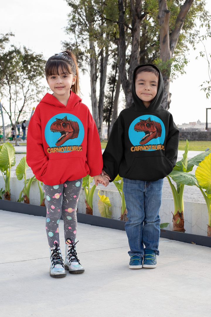 Two children stand outdoors hand in hand, both dressed in Jurassic Studio Kids Carnotaurus Head Hoodies—one girl in red with patterned leggings, one boy in black paired with jeans. Lush green plants form the backdrop.
