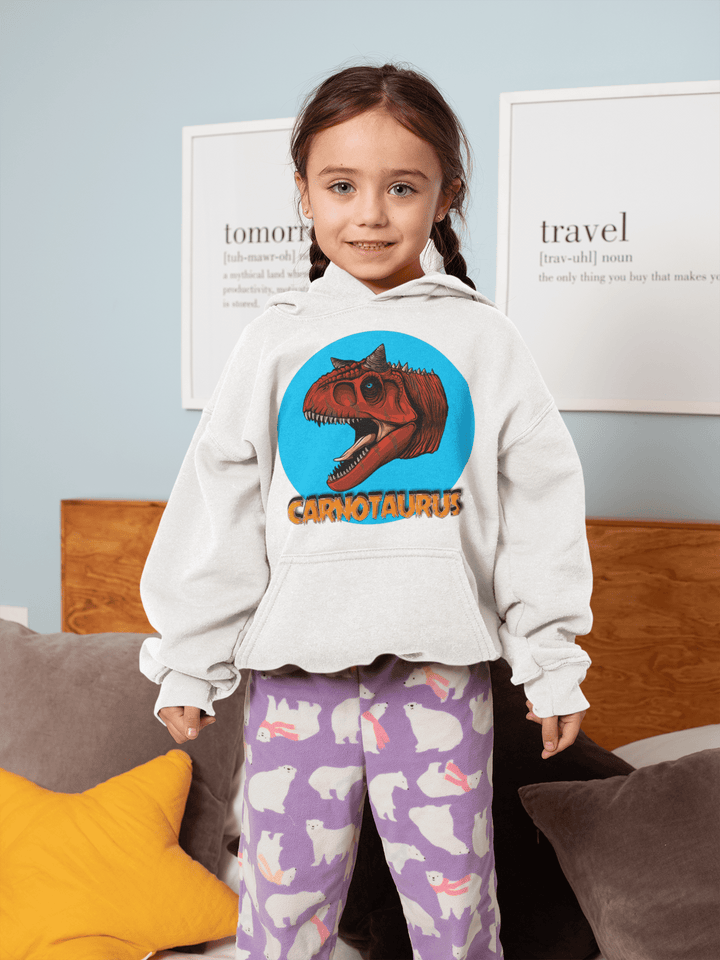 A young girl smiles on a bed wearing the Jurassic Studio Kids Carnotaurus Head Hoodie—a white hoodie with a red dinosaur head and CARNOTAURUS text—paired with purple bunny-print pajama pants. Decorative pillows and framed art are behind her.