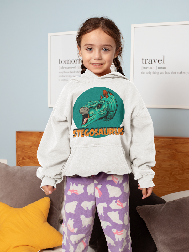 A young girl with long brown hair stands on a bed, smiling and wearing the Jurassic Studio Kids Stegosaurus Head Hoodie along with purple pajama pants featuring white bunny prints.