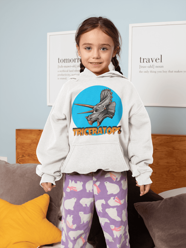 A young girl smiles on a bed, wearing the Jurassic Studio Kids Triceratops Head Hoodie with purple pajama pants featuring white bunnies. Pillows and wall art decorate the background.