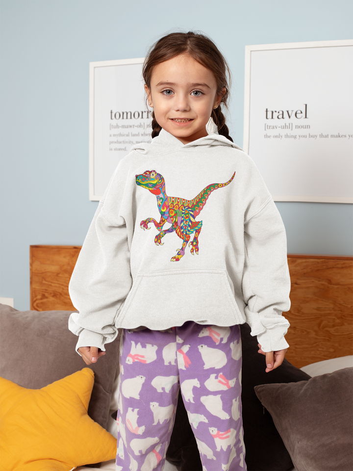 A young girl stands on a bed wearing a white Jurassic Studio Kids Velociraptor Zentangle Hoodie with a colorful dinosaur design and purple pajama pants with white bunny patterns, smiling at the camera with pillows and framed wall art behind her.