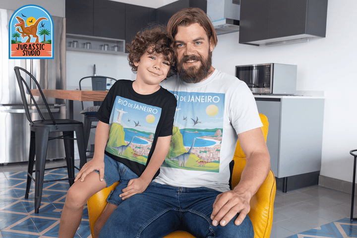 A smiling man and child sit in a modern kitchen, both wearing matching Men Rio de Janeiro T-Shirts from Jurassic Studio—a great dinosaur fan gift. The Jurassic Studio logo appears as an exclusive mark in the images top left corner.