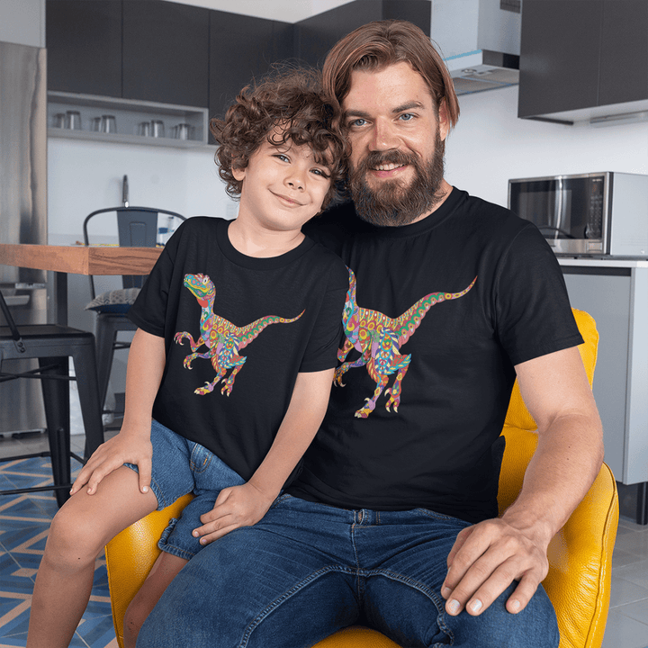 A smiling man and child sit together in a kitchen, both wearing black Jurassic Studio Men Velociraptor Zentangle T-Shirts with a colorful dinosaur design. The child is on the mans lap, and they look happy and relaxed.