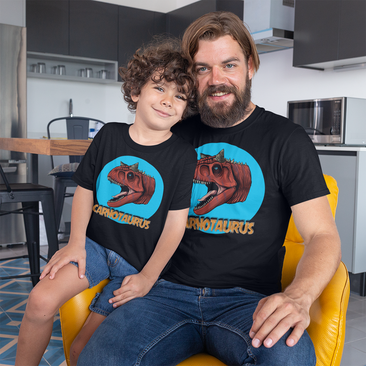 A man and a boy smile together in a modern kitchen, both wearing Jurassic Studio’s Men Carnotaurus Head T-Shirt. The boy sits on the mans lap on a yellow chair—an ideal dinosaur fan gift.