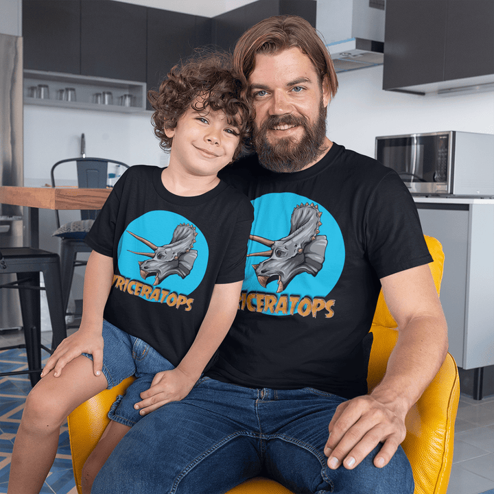 A smiling man and child sit together in a kitchen, both wearing matching Jurassic Studio Men Triceratops Head T-Shirts—perfect for dinosaur fans, featuring a bold triceratops graphic with TRICERATOPS printed below.