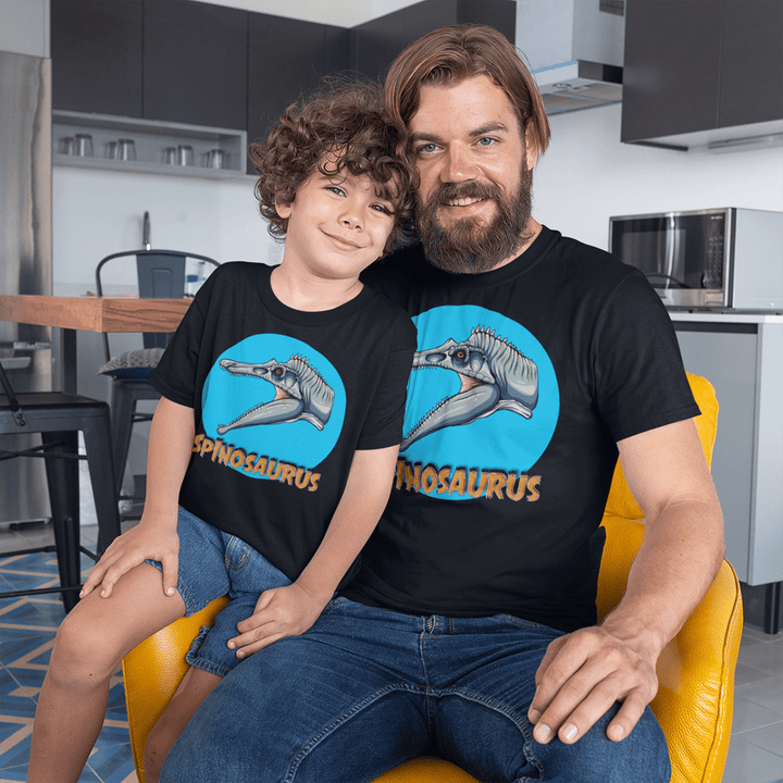 A smiling man and young boy relax in a modern kitchen, both wearing the Jurassic Studio Men Spinosaurus Head T-Shirt—a perfect, colorful dinosaur fan gift and exclusive tee for their joyful moment together.