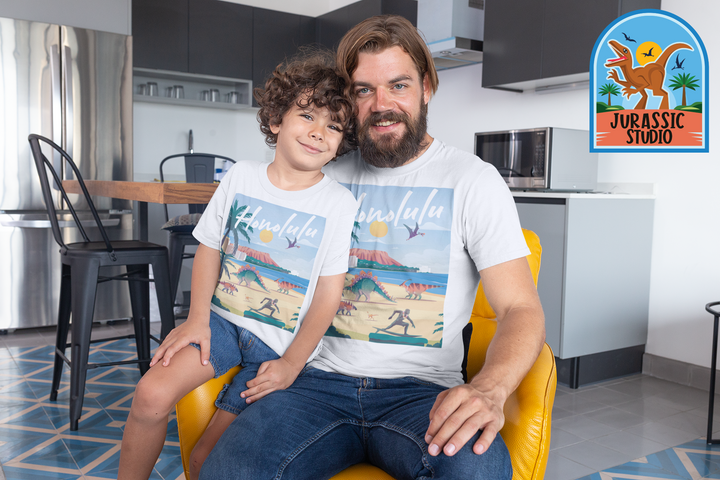 A smiling man and child sit in a modern kitchen, both wearing matching Men Honolulu T-Shirts by Jurassic Studio, featuring a beach scene and dinosaur logo—an ideal gift for any dinosaur fan.