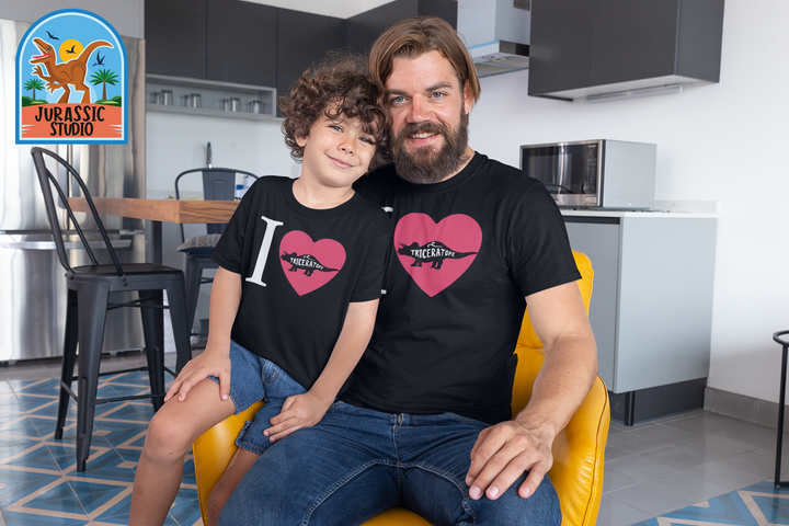 A man and boy smile in a sleek kitchen, both wearing Men I Love Triceratops T-Shirts by Jurassic Studio. The dinosaur-themed logo appears at top left—a perfect gift for dino fans and a Jurassic Studio exclusive.
