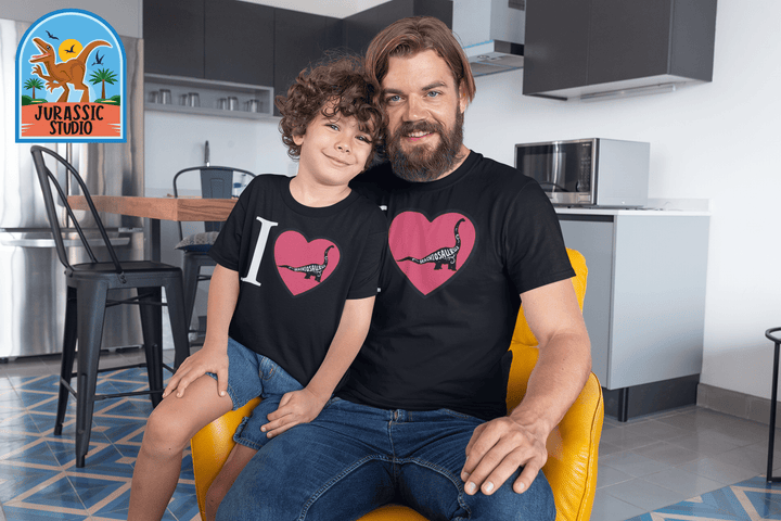 A smiling man and boy sit together in a kitchen wearing matching Men I Love Brachiosaurus T-Shirts from Jurassic Studio. The Jurassic Studio logo in the top left marks this as the perfect gift for dinosaur fans and an exclusive from the brand.