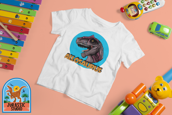 The Toddler Allosaurus Head T-Shirt by Jurassic Studio features a bold dinosaur graphic and text, ideal for young dino fans. Displayed among colorful toys on a peach background, with the Jurassic Studio logo in the bottom left corner.